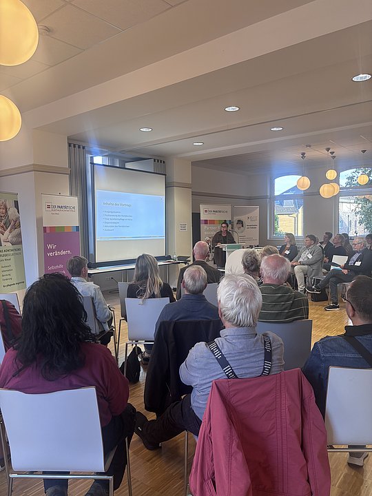 Foto vom gesamten Raum. Sitzende Menschen. Im Hintergrund Leinwand mit Präsentation